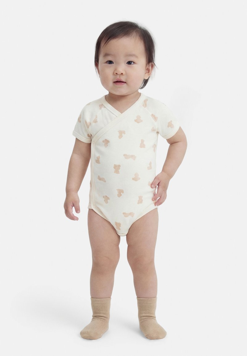 Μικρό παιδί που στέκεται, φορώντας κρεμ φορμάκι με ανοιχτά καφέ ζωάκια και μπεζ κάλτσες, με φόντο απλό λευκό τοίχο.