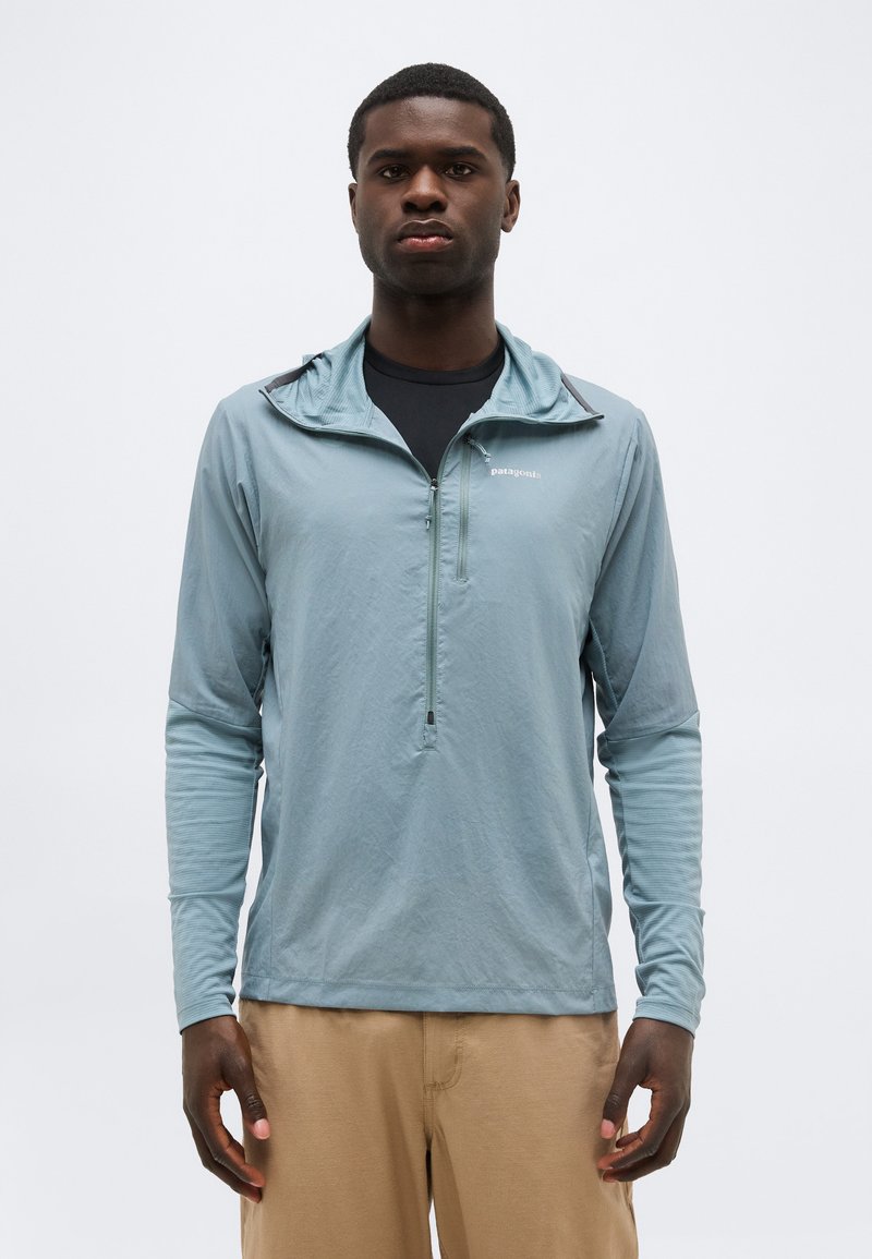 Άνδρας με ανοιχτό μπλε μπουφάν Patagonia με φερμουάρ στο ένα τέταρτο, πάνω από μαύρο πουκάμισο και μπεζ παντελόνι, στέκεται μπροστά σε απλό λευκό φόντο.