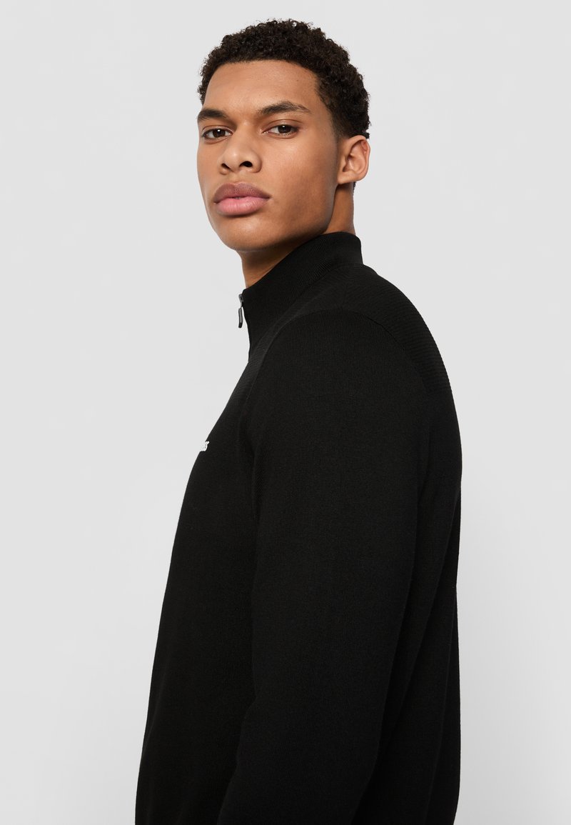 Schwarzer Pullover mit Reißverschluss am Hals, eng anliegendem Design und geripptem Material, mit einem dezenten Logo auf der Brust und einer glatten Oberfläche.