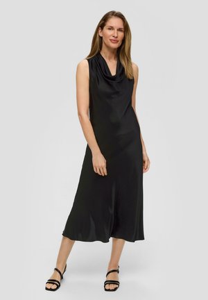 Zwarte mouwloze jurk met een loeihals, een vloeiende silhoët en een gladde, glanzende stof. Gepaard met zwarte sandaaltjes met banden.