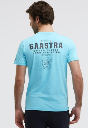 Mann mit braunen Haaren trägt ein hellblaues Kurzarmhemd mit großem schwarzem "GAASTRA"-Schriftzug und segelthematischem Text auf der Rückseite, steht vor einem einfarbigen Hintergrund.