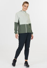 Letvægts, tofarvet jakke i lys grøn og mørk grøn, med lynlås og hætte. Kombinationen er med sorte tapered bukser og sorte sko.