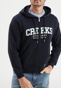 Felpa con zip di colore blu navy, realizzata in misto cotone, con tasca frontale e lettering a contrasto bianco e azzurro chiaro sul petto.
