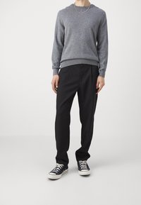 Mężczyzna w szarej długiej koszulce, czarnych spodniach, czarno-białych sneakersach i srebrnym łańcuszku, stojący na białym tle.