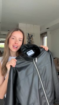 Γυναίκα με μακριά μαλλιά κρατά ένα μαύρο δερμάτινο μπουφάν με μπροστινό φερμουάρ μέσα σε ένα μοντέρνο σαλόνι με έργα τέχνης στον τοίχο.