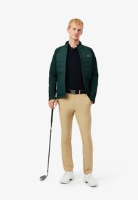 Donkergroene gewatteerde jas over een zwarte polo, beige broek, met een golfclub in de hand, witte schoenen. Bevat Lacoste branding op de jas.
