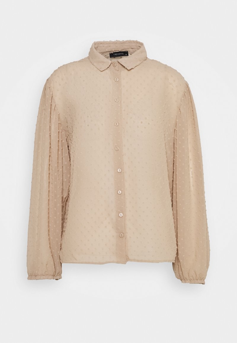 Blouse beige texturée avec devant boutonné, col et manches longues froncées. Le tissu présente des motifs à points en relief pour plus de détails.