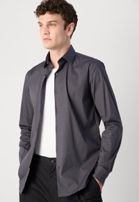 Graues Langarmhemd mit strukturiertem Kragen, geknöpften Vorderteil und glatter Textur, über einem weißen T-Shirt getragen und mit dunklen Hosen kombiniert.
