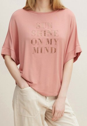 Femme portant un haut rose ample avec le texte « SUNSHINE ON MY MIND » en lettres brillantes, associé à un pantalon beige clair, main dans la poche.