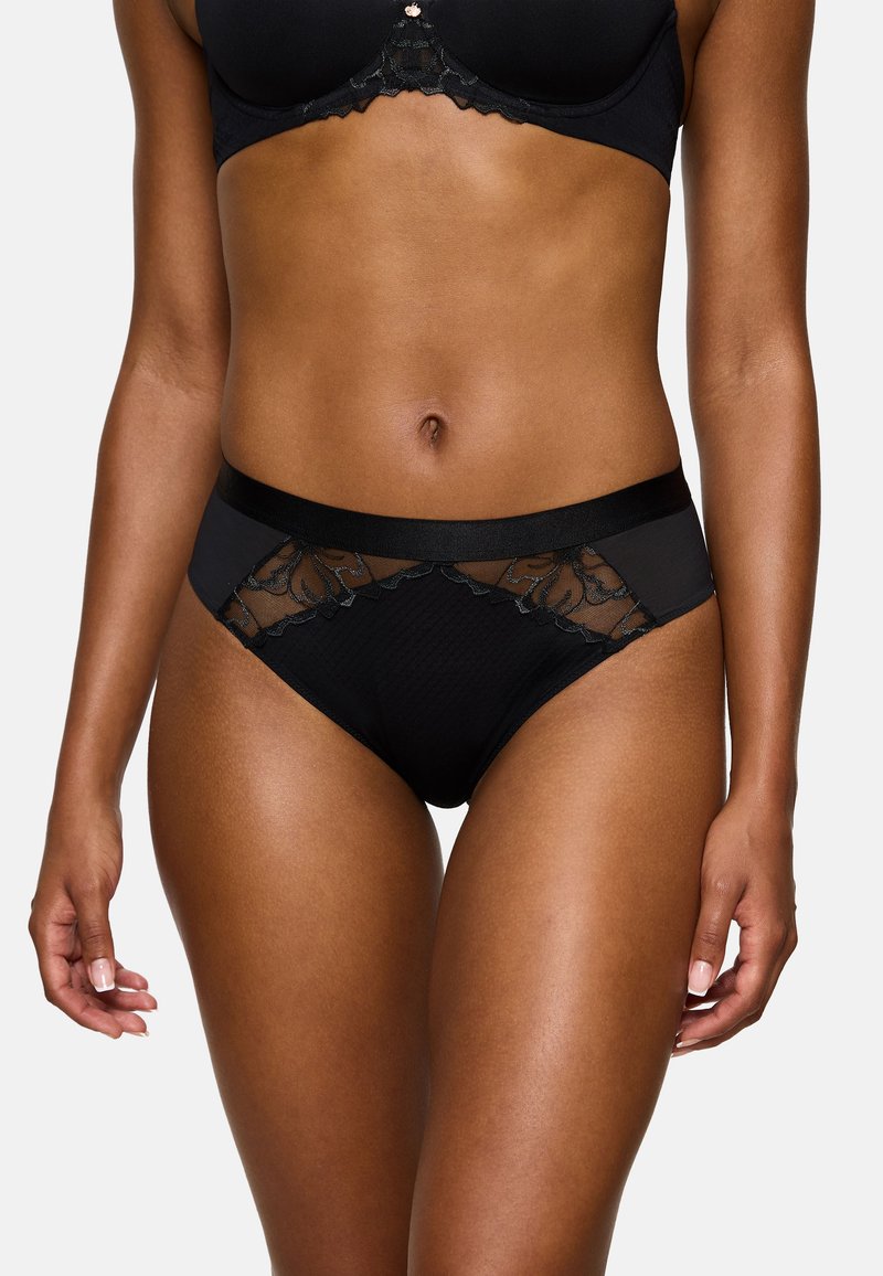 Vrouw in zwart beha met kanten rand en bijpassende slip, staand met ontspannen armen langs het lichaam tegen een effen achtergrond.