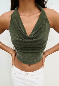 Olivegrön topp med hög krage, tillverkad av texturerat material; åtsittande design med rynkade detaljer. Kombinerad med vita denimbukser.