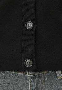 Cardigan en maille noire avec texture côtelée et trois grands boutons ronds. Associé à un pantalon en denim gris avec des coutures jaunes.