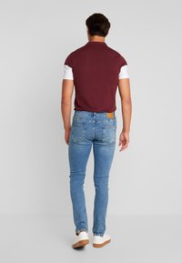 Vinröd pikétröja med vita ärmar, blå jeans i skinny fit och vita sneakers; sedd bakifrån mot en neutral bakgrund.