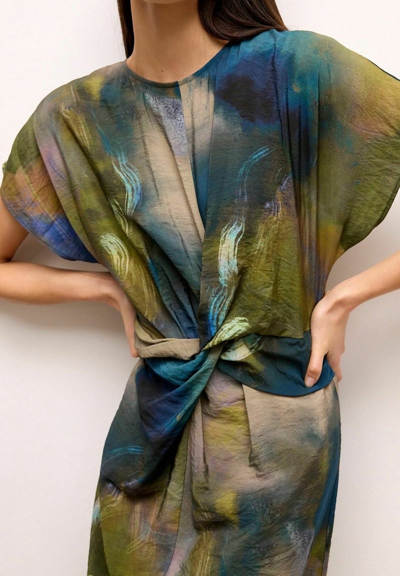 Femme portant une robe ample à manches courtes avec un motif abstrait bleu, vert et beige, présentant un détail de nœud torsadé à l'avant.