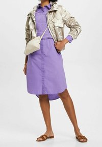 Metallic silverjacka, lila skjortklänning med texturerad liv, och ljus beige crossbody-väska. Bruna sandaler fulländar outfiten.