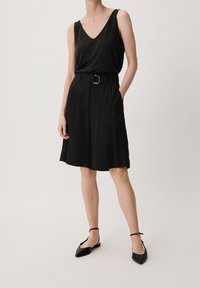 Haut sans manches noir avec un décolleté en V associé à une jupe noire arrivant au genou avec une ceinture. Le modèle porte des ballerines noires à bouts pointus. Texture lisse.