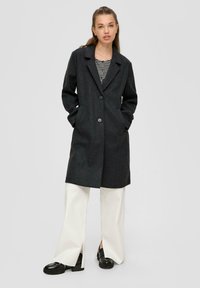 Dunkelgrauer Wollmantel mit Reverskragen, knöpfbarer Einknopfleiste und seitlichen Taschen. Kombiniert mit weißen Hosen mit gesplittetem Saum und schwarzen Schuhen.