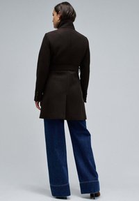 Abrigo de lana marrón con cuello alto, cintura ceñida con cinturón y puños abotonados. Combinado con unos vaqueros de pierna ancha azul con un dobladillo cosido.