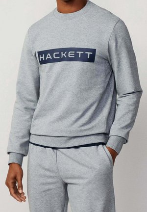 Mies, jolla on vaaleanharmaa collegepaita ja siihen sopivat collegehousut, paidassa rintakehän kohdalla laivastonsininen suorakulmio, jossa lukee "HACKETT".