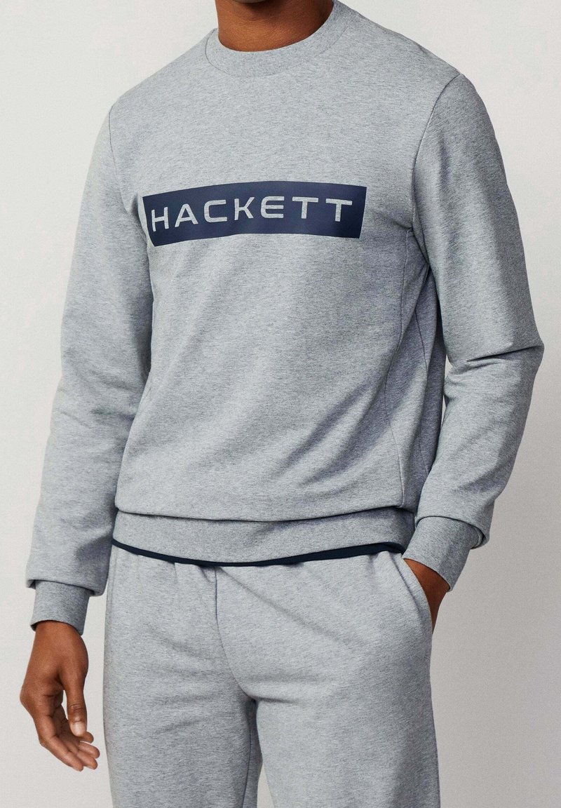 Homme portant un sweat-shirt gris clair et un pantalon de survêtement assorti, le sweat-shirt affichant le texte "HACKETT" dans un rectangle bleu marine sur la poitrine.