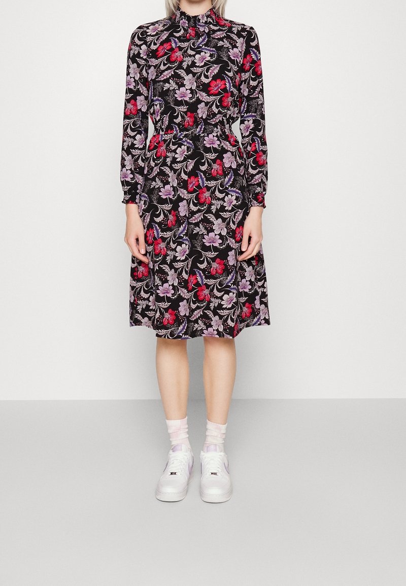 Femme portant une robe noire longueur genou avec un motif floral rouge et violet, manches longues, et des baskets blanches avec des chaussettes rose clair.