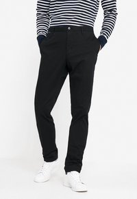 Chinos neri con una texture liscia, dotati di chiusura a bottone e tasche laterali. Abbinati a una camicia a maniche lunghe a righe blu navy e sneakers bianche.