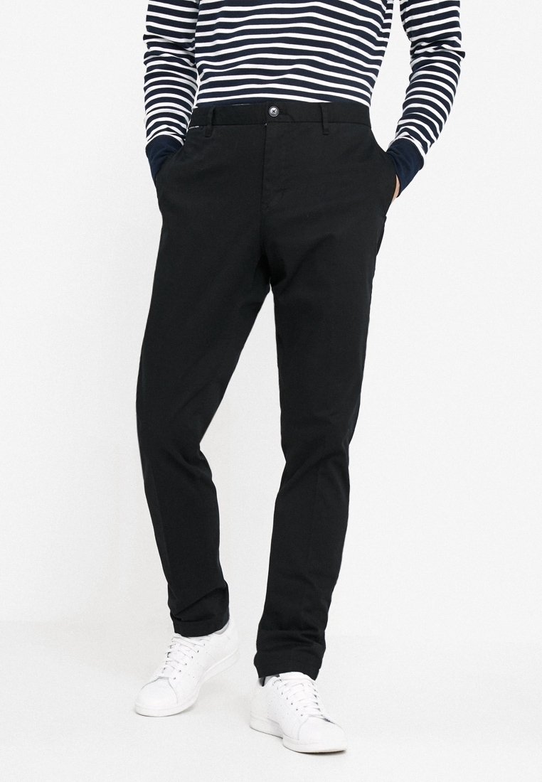 Chinos neri con una texture liscia, dotati di chiusura a bottone e tasche laterali. Abbinati a una camicia a maniche lunghe a righe blu navy e sneakers bianche.
