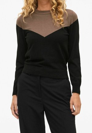 Femme portant un pull à manches longues noir et marron avec un motif chevron, associé à un pantalon noir, debout devant un fond uni.