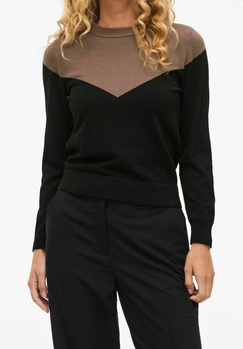 Femme portant un pull à manches longues noir et marron avec un motif chevron, associé à un pantalon noir, debout devant un fond uni.