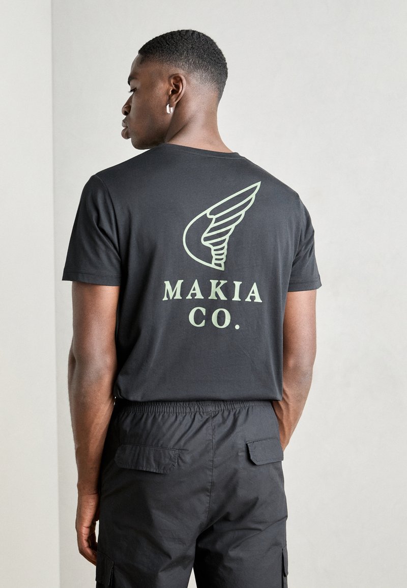 Black short-sleeve t-shirt featuring a large green wing graphic and "MAKIA CO." text on the back. Fabric appears smooth and lightweight.
