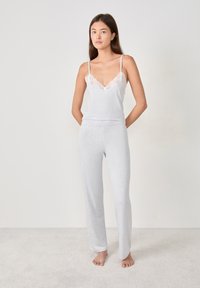 Jonge vrouw die blootvoets staat in een witte camisole met kantafwerking en bijpassende losse broek op een lichte tapijt vloer.