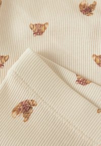 Geribbelde stof in crème kleur met een herhaald croissantpatroon in bruin. Het materiaal heeft een lichte textuur met zichtbare stiksels.