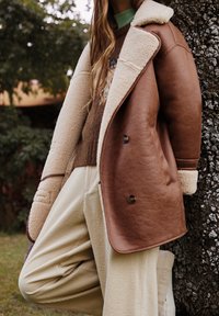 Brun läderjacka med krämfärgad fuskpäls vid kragen och ärmarna, dubbelknäppt design, kombinerad med krämfärgade kordflorsbyxor.