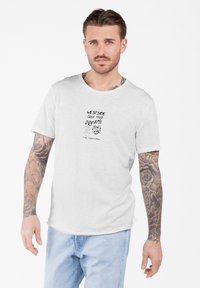 Hombre con tatuajes en ambos brazos, vestido con una camiseta blanca con texto negro y unos jeans azul claro, de pie contra un fondo blanco.