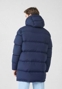 Homme portant un manteau d'hiver matelassé bleu marine avec une grande capuche, se tenant de dos devant un fond clair.