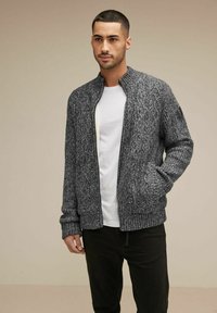 Grå stickad bomberjacka med ribbade muddar och nederkant, med dragkedja framtill och sidofickor, bärs över en vit t-shirt och svarta byxor.