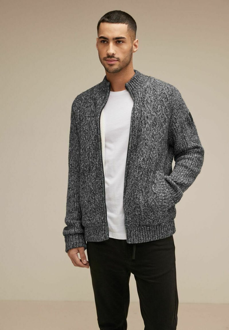 Grå stickad bomberjacka med ribbade muddar och nederkant, med dragkedja framtill och sidofickor, bärs över en vit t-shirt och svarta byxor.