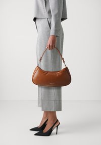 Borsa a spalla in pelle marrone con forma curva e corta tracolla. Indossata con un outfit a quadri bianco e nero e tacchi a stiletto neri.