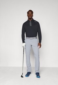 Muž v čiernej polokošeli s dlhým rukávom, šedých nohaviciach a modrých golfových topánkach, ktorý v jednej ruke drží golfovú palicu a v druhej ruke golfovú loptičku.