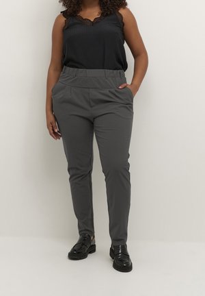 Frau mit lockigem Haar trägt ein schwarzes ärmelloses Spitzenoberteil, graue, schmal zulaufende Hosen mit elastischem Bund und schwarze Loafers, steht vor einer beigen Wand.