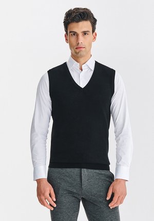 Czarny dzianinowy kamizelka w serek na białej koszuli z kołnierzykiem, zestawiona z szarymi spodniami. Gładka tekstura, dopasowany krój, minimalistyczne detale.