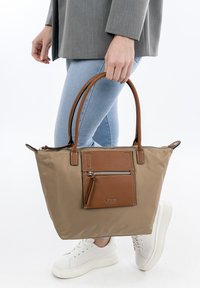Borsa tote in nylon beige con dettagli in pelle marrone, dotata di una tasca con cerniera frontale e doppio manico, mostrata da una persona in jeans azzurri.