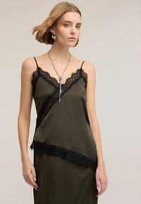 Camisole di raso verde oliva con bordatura in pizzo nero, caratterizzata da un design diagonale e spalline sottili. Modella con una collana d'argento.
