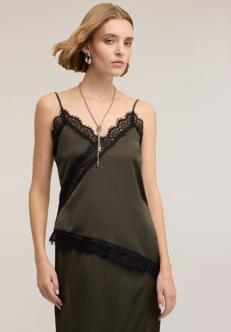 Camisole di raso verde oliva con bordatura in pizzo nero, caratterizzata da un design diagonale e spalline sottili. Modella con una collana d'argento.
