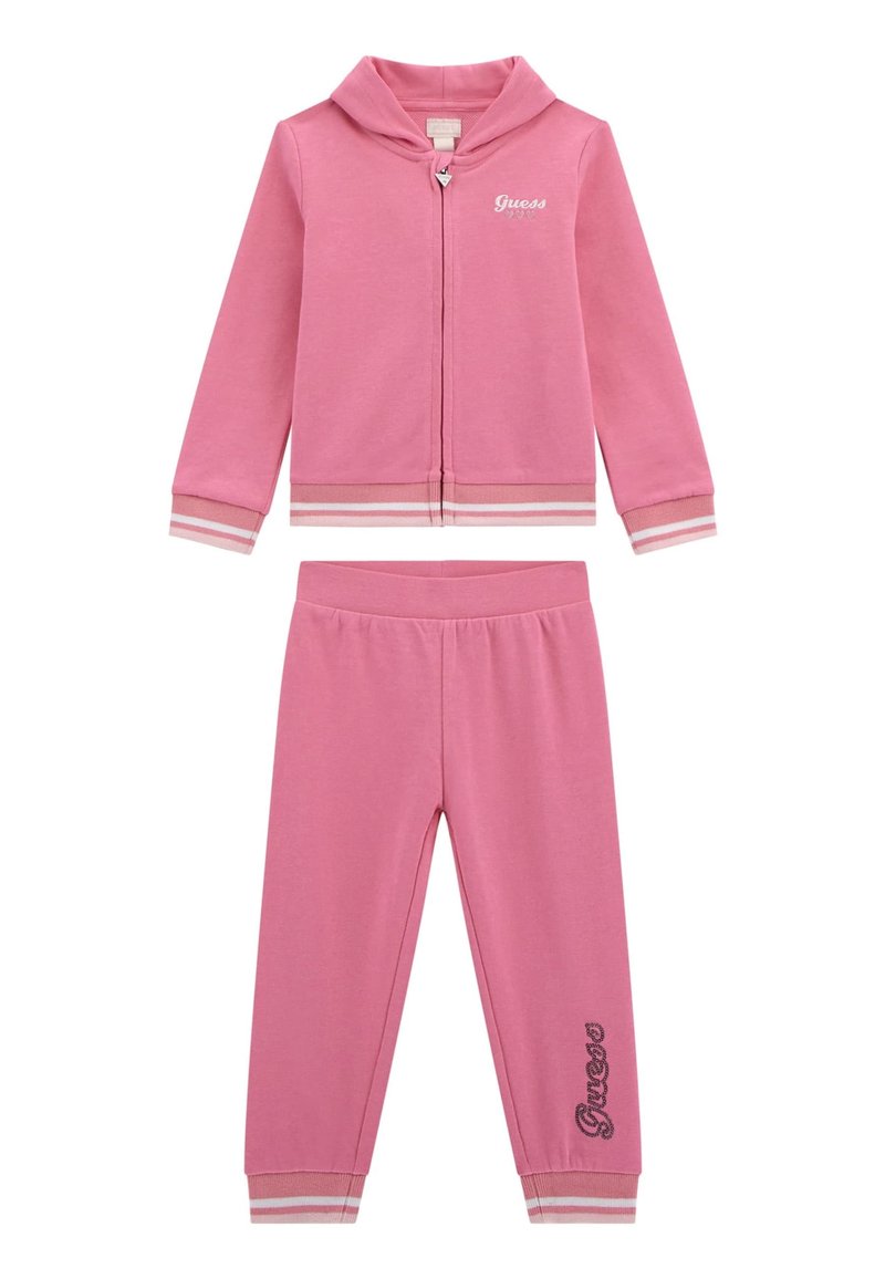 Ensemble en sweat-shirt et pantalon rose en matériau doux. Comprend un hoodie zippé, des poignets rayés, et le logo "Guess" sur la veste et le pantalon.