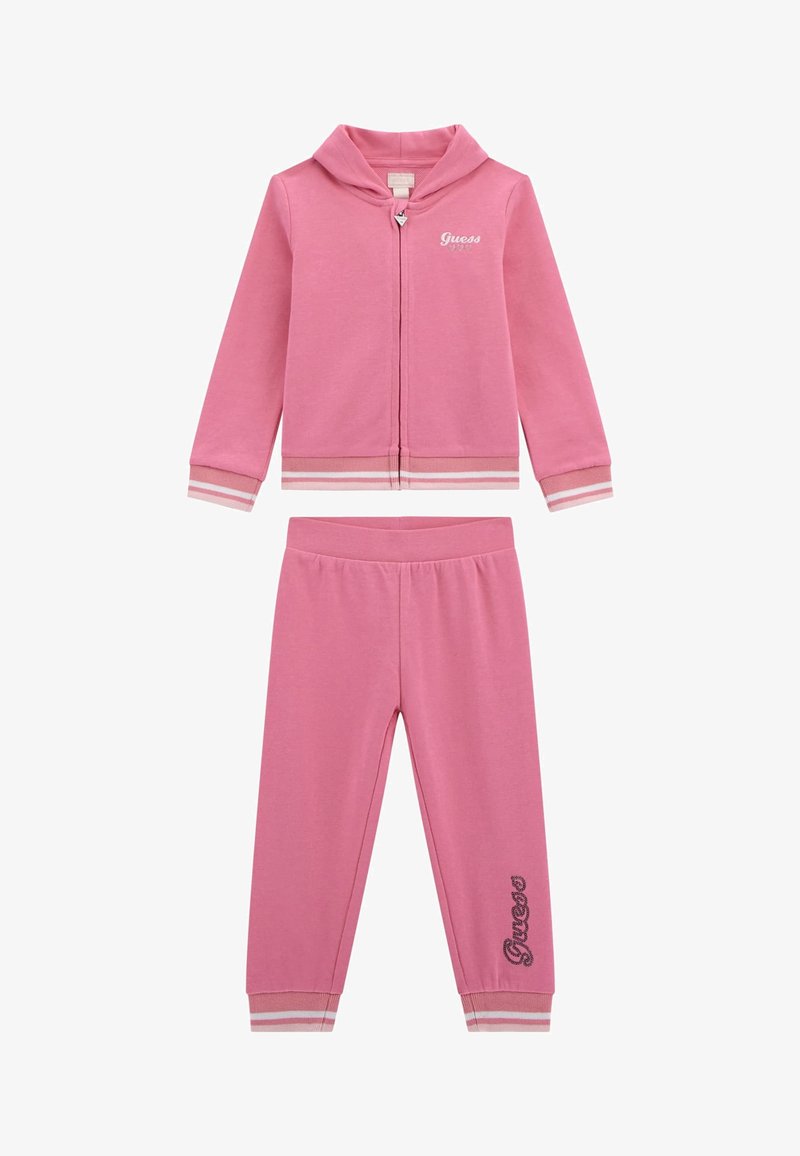 Set di felpa e pantaloni rosa realizzato in materiale morbido. Presenta una felpa con zip, polsini a righe e il logo "Guess" sulla giacca e sui pantaloni.