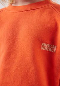 Krupni plan narančastog pletivnog džempera s ispisanim tekstom "AMERICAN VINTAGE" na prsima, blizu ramena.