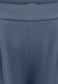 Blå teksturerte bukser med høy midje, bred linning og liten svart etikett med teksten "ONLY CARMAKOMA" midt på baksiden.