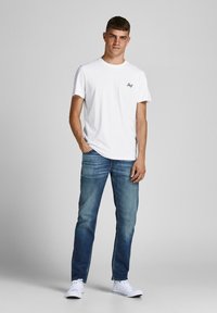 Weißes Baumwoll-T-Shirt mit einem kleinen schwarzen Logo auf der Brust, kombiniert mit blauen Jeans und weißen Sneakers. Lässiger Schnitt, keine Muster.