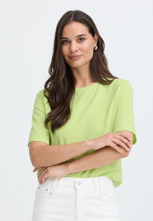 Femme aux longs cheveux bruns, portant un t-shirt vert clair à manches courtes et un pantalon blanc, debout, les bras croisés, sur un fond uni.